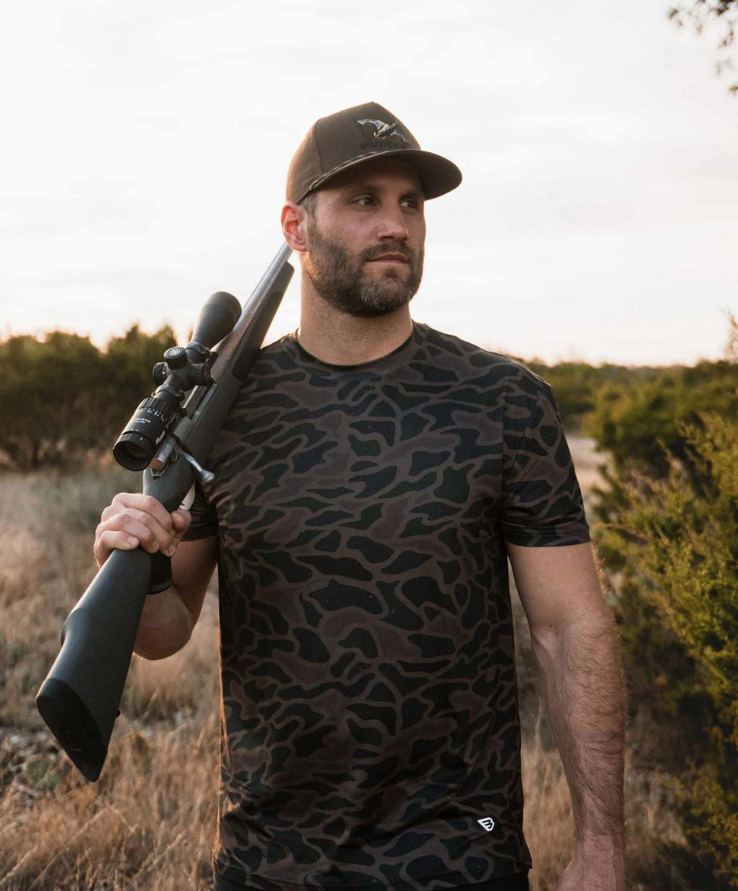 A man wearing a dark brown and black camouflage t-shirt and a matching baseball cap, holding a rifle with a scope over his shoulder in an outdoor field setting.