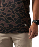 Close-up of a person wearing a dark camouflage-patterned t-shirt and beige pants, with a black smartwatch on the wrist.