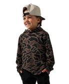 A young boy wearing a brown and black camouflage patterned hoodie, black pants, and a light beige baseball cap, shown against a plain white background.