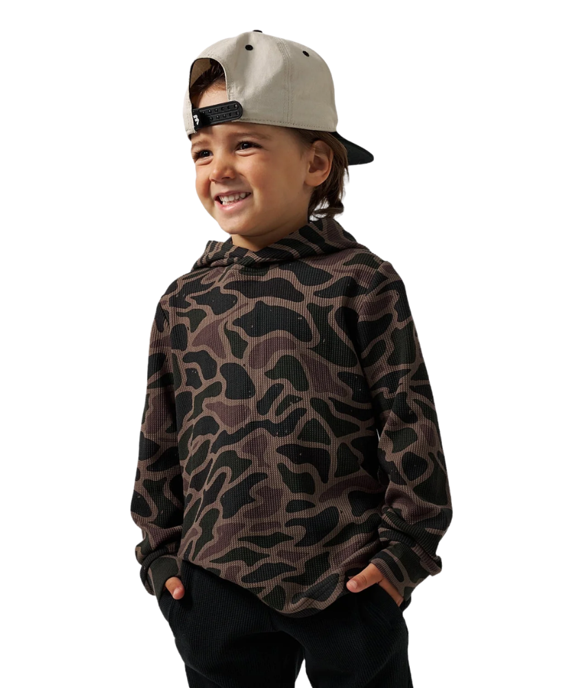 A young boy wearing a brown and black camouflage patterned hoodie, black pants, and a light beige baseball cap, shown against a plain white background.