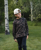 A child standing outdoors in a grassy park wearing a dark camouflage hoodie, dark pants, and a white baseball cap with a graphic print.