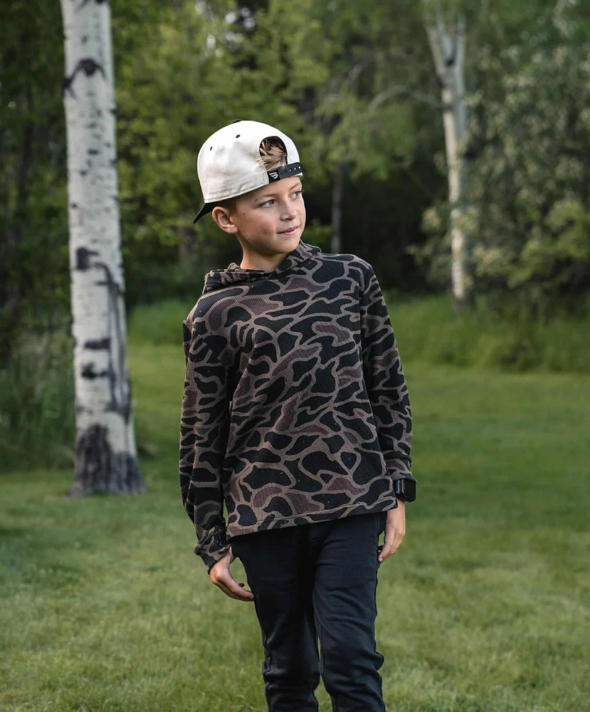 A child standing outdoors in a grassy park wearing a dark camouflage hoodie, dark pants, and a white baseball cap with a graphic print.