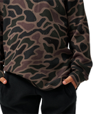 A close-up of a person wearing a long-sleeve waffle-knit top with a dark camouflage pattern in black, olive green, and brown, paired with black textured pants.
