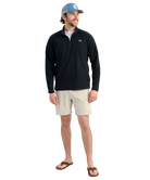 Man wearing a black long-sleeve HUK PULLOVER with beige shorts, and a blue cap on a white background