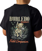 A child wearing a black short-sleeve T-shirt, shown from the back. The shirt features a large graphic print with the words 'BURLEBO Outdoors' and 'Field Companion' in orange and beige lettering, surrounding an illustration of a yellow Labrador retriever holding a fish in a marsh.