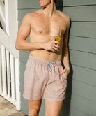 Man wearing striped swim shorts holding a drink against a gray wall.