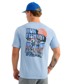 Man wearing a light blue HUK t-shirt with a dog graphic design on the back, standing against a white background.