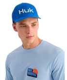 Man wearing a light blue t-shirt with Huk logo and a matching blue cap on a white background