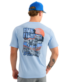 Man wearing a light blue HUK t-shirt with a dog graphic design on the back, standing against a white background.