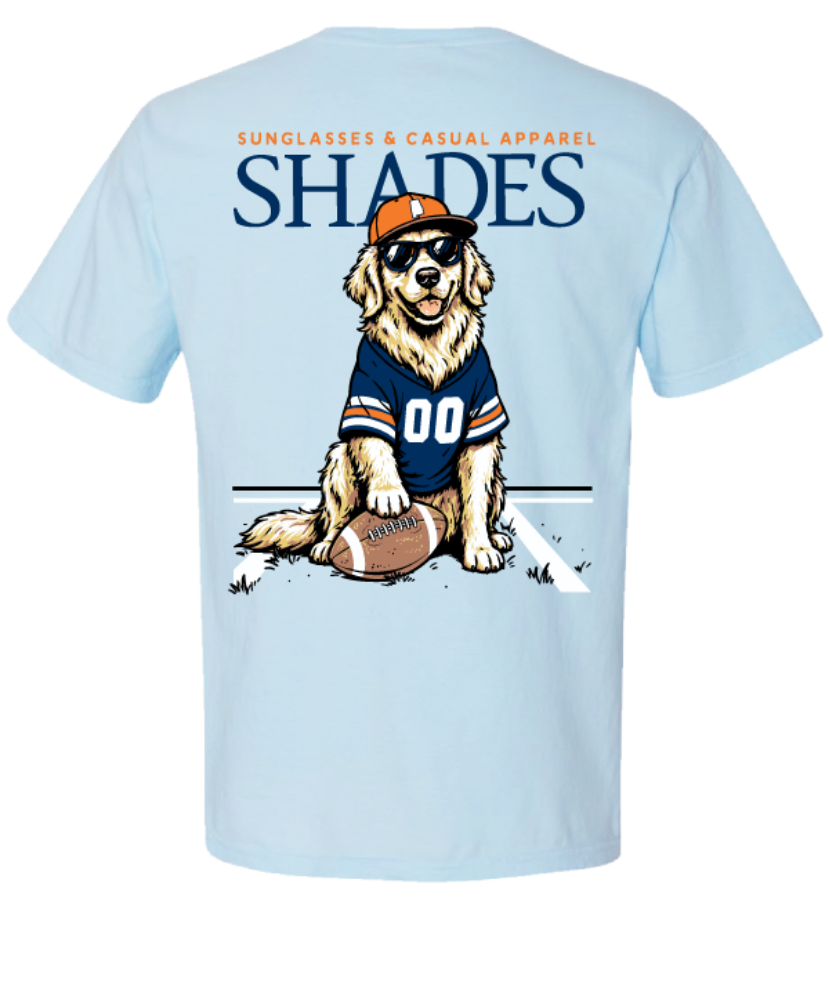 A light blue short-sleeve t-shirt shown from the back, featuring a graphic of a golden retriever wearing a navy and orange football jersey, sunglasses, and an orange cap, sitting next to a football. The word 'SHADES' is printed in large navy letters above the dog, with smaller orange text reading 'SUNGLASSES & CASUAL APPAREL' above that.