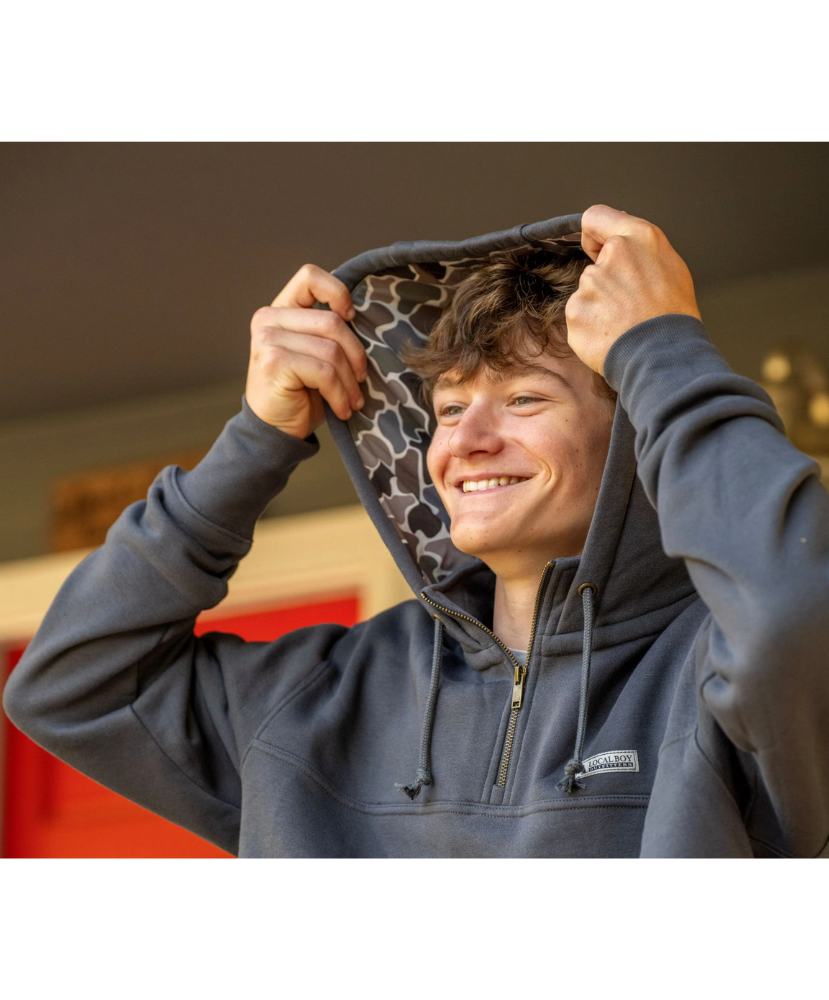 A smiling man wearing a dark gray hoodie with a patterned interior hood lining, holding the hood up with both hands.
