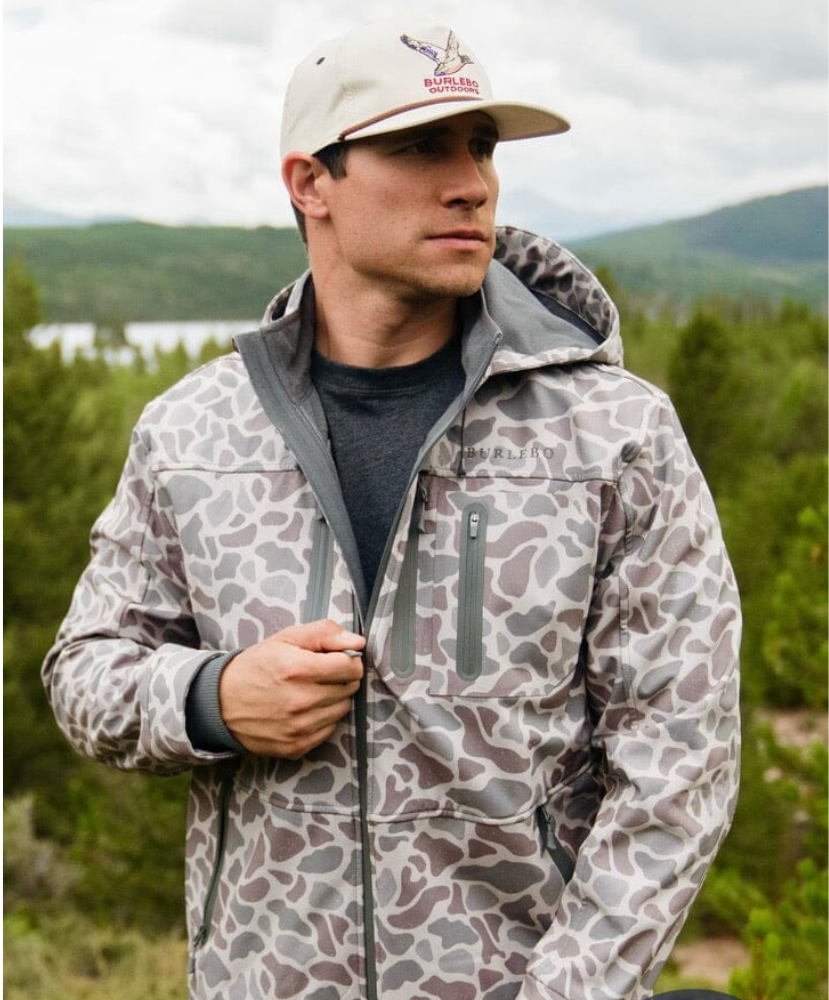 Man wearing a patterned jacket and cap outdoors with a lake and mountains in the background