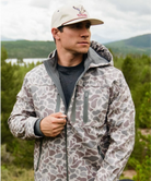 Man wearing a patterned jacket and cap outdoors with a lake and mountains in the background