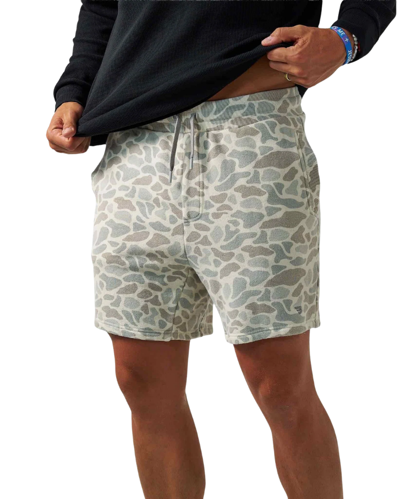 A person wearing patterned shorts and pulling up a black long-sleeve shirt against a plain white background.