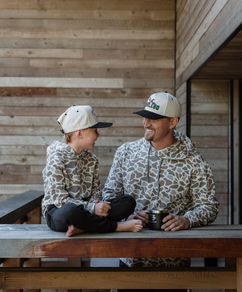 A man and a child sitting on a wooden deck, both wearing matching beige and brown animal-print hoodies and matching baseball caps with black brims.