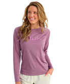 A woman wearing a long-sleeve purple Nuk Fishing shirt with a pink logo on the chest, paired with light beige pants, set against a plain white background.