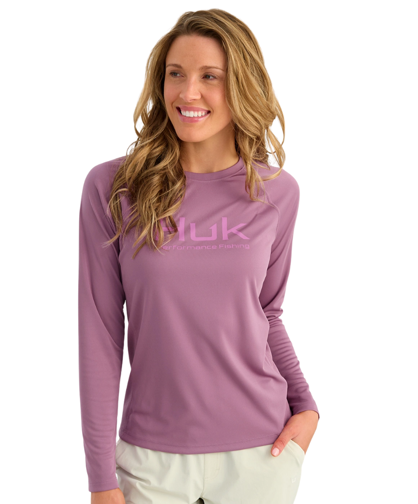 A woman wearing a long-sleeve purple Nuk Fishing shirt with a pink logo on the chest, paired with light beige pants, set against a plain white background.