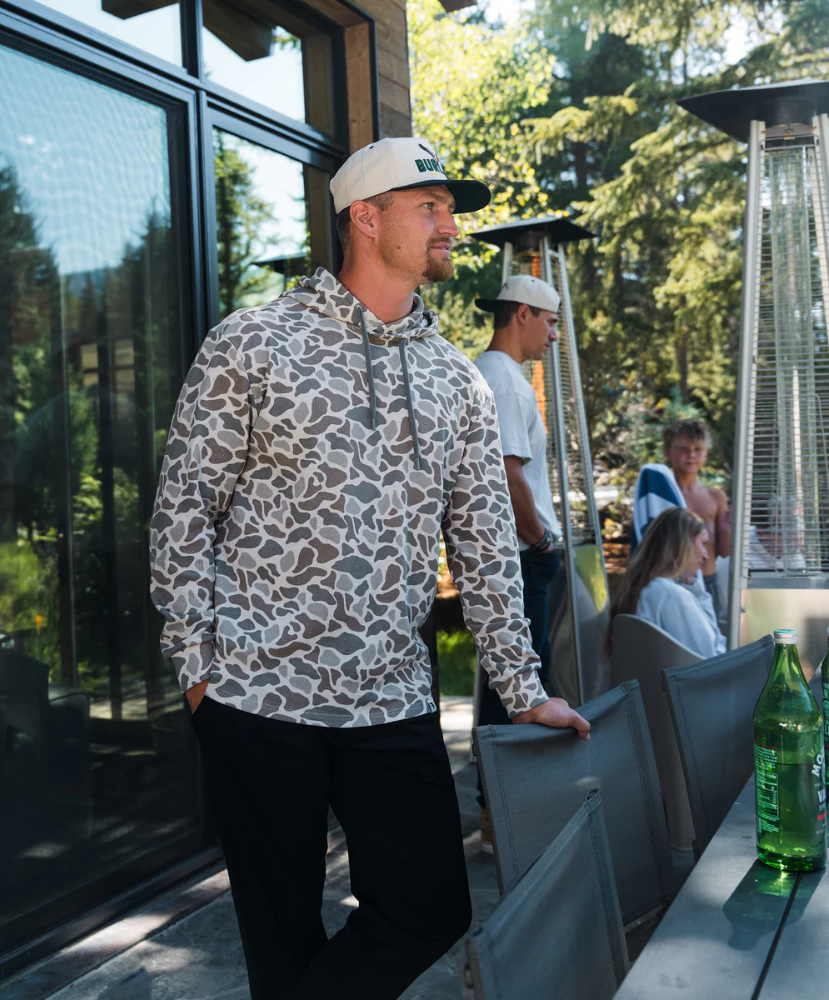 Man wearing a THERMAL hoodie and cap standing outdoors with trees and people in the background