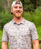 Man wearing a patterned polo shirt and cap outdoors with greenery in the background