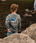 Child wearing a gray long sleeve t-shirt with a yellow lab dog graphic and text, standing near rocks and water.