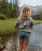 Girl wearing a gray t-shirt with a duck stamp design and 'Burlebo Outdoors' branding, standing by a water body with mountains in the background.