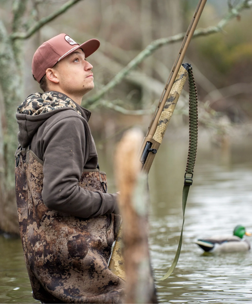 A man in camouflage hunting waders and a brown hoodie stands in shallow water holding a rifle with a camouflage stock and green sling strap. A mallard duck is visible in the background water, with blurred tree branches behind him.