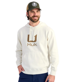 Man wearing a cream hoodie with 'Huk' logo and a cap on a white background