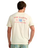 Man wearing a cream HUK t-shirt with red MARINA HUK and graphics on the back, standing against a white background.