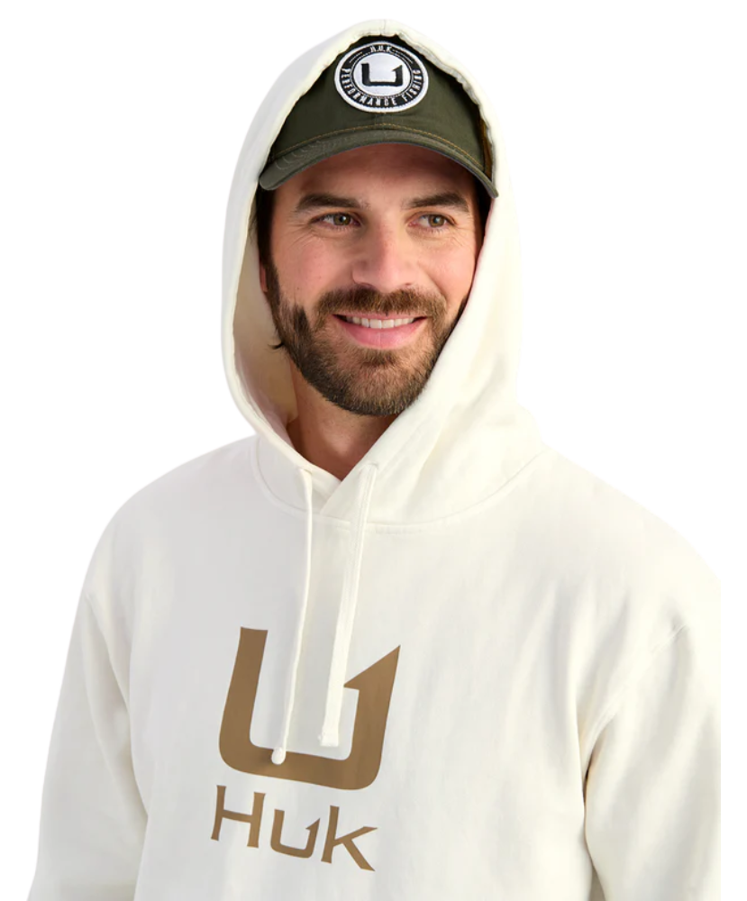 Man wearing a cream hoodie with 'Huk' logo and a cap on a white background