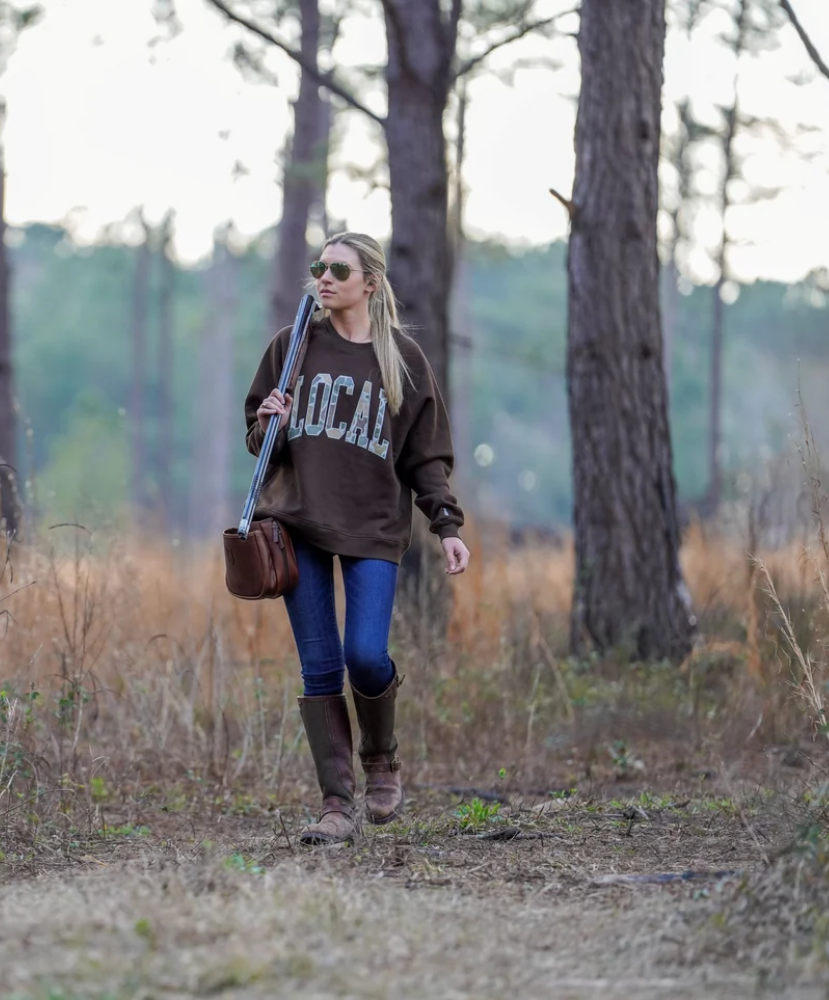 A woman walking on a forest path wearing a brown oversized sweatshirt with 'LOCAL' printed across the chest, blue jeans, brown boots, and carrying a brown shoulder bag.