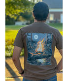 Person wearing a DIRTY MARSH brown t-shirt with a graphic design of a campfire and moon on the back, standing outdoors.