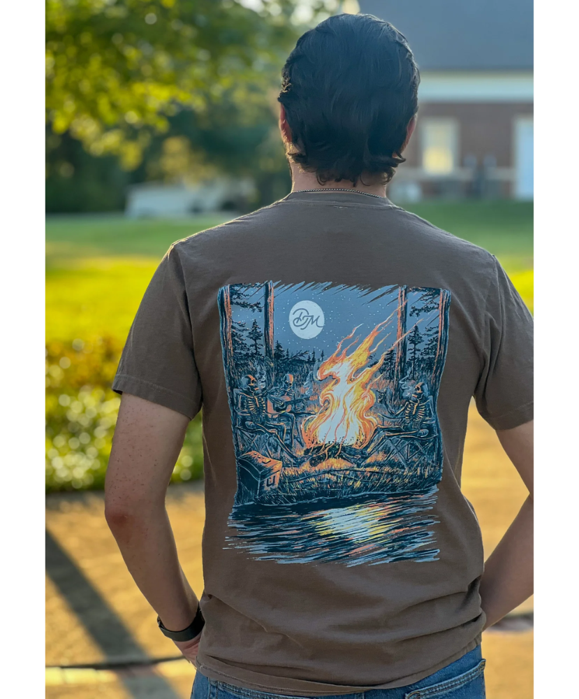 Person wearing a DIRTY MARSH brown t-shirt with a graphic design of a campfire and moon on the back, standing outdoors.