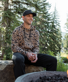 Man wearing a Camoflage print hoodie and black cap sitting outdoors with trees in the background
