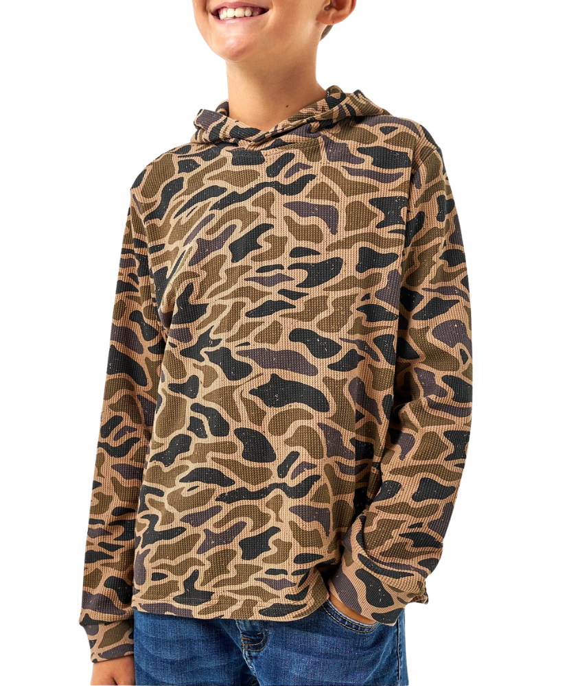 A child wearing a long-sleeve hooded shirt with an allover camouflage-style pattern in tan, brown, and black, paired with blue jeans against a plain white background.