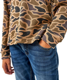 A close-up of a person wearing a waffle-knit long-sleeve top with a tan, brown, and black abstract camouflage pattern, paired with blue jeans.