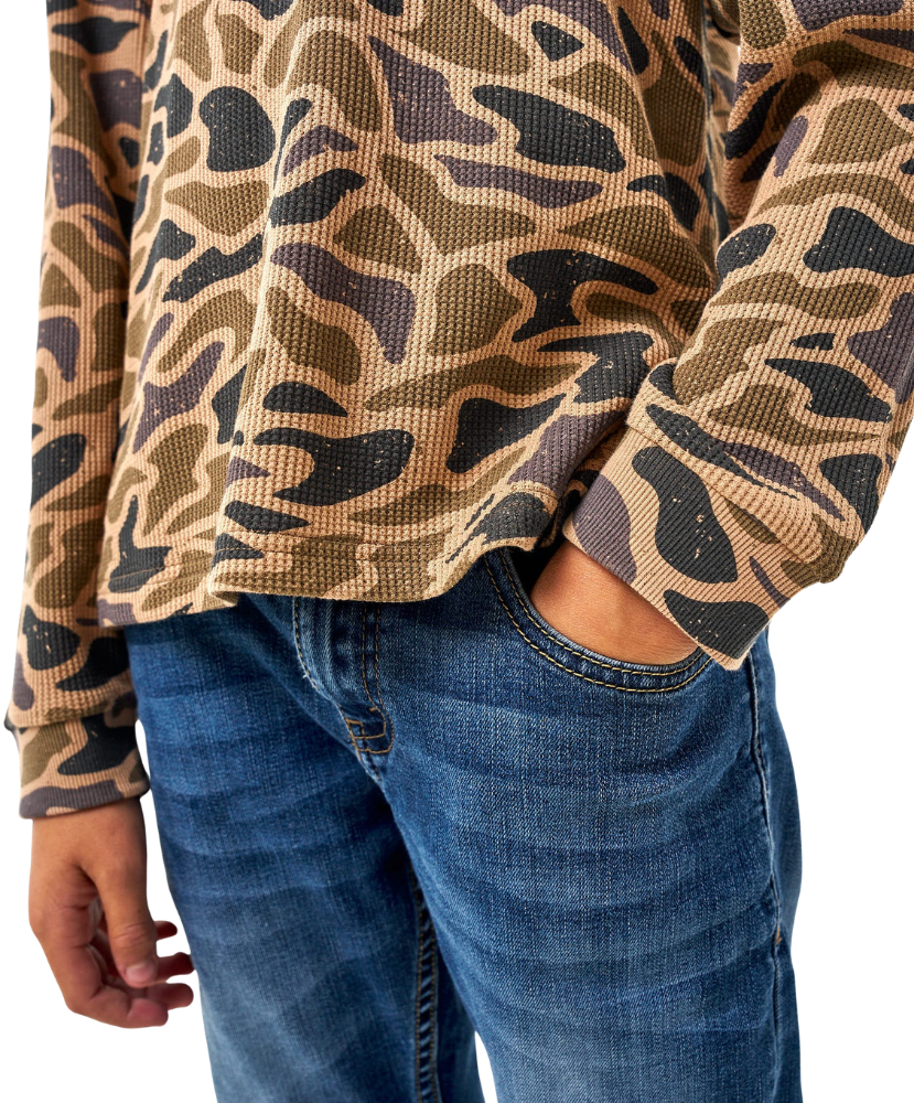 A close-up of a person wearing a waffle-knit long-sleeve top with a tan, brown, and black abstract camouflage pattern, paired with blue jeans.