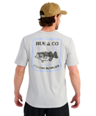 Man wearing a gray t-shirt with Huk & Co fishing supplies logo on the back, Bass graphic, standing against a white background.