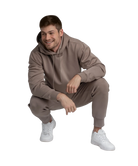 Man wearing a brown hoodie and jogger pants with white sneakers on a white background