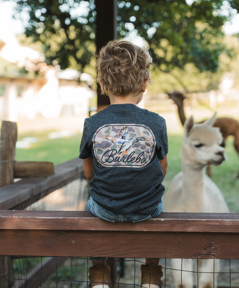 Child wearing a shirt with 'Burlebo' on it, sitting by a fence looking at animals.