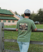 Man wearing a green t-shirt with a graphic design, standing by a wooden fence in an outdoor setting.