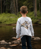 Child standing by a forest stream wearing a gray long sleeve t-shirt with a mallard duck design.
