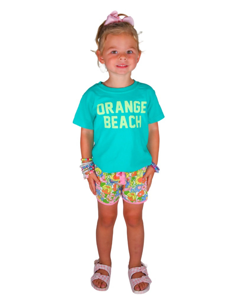 Young girl wearing a turquoise 'Orange Beach' shirt and colorful shorts on a white background