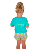 Child wearing a turquoise shirt with 'SHADES' branding and floral shorts on a white background