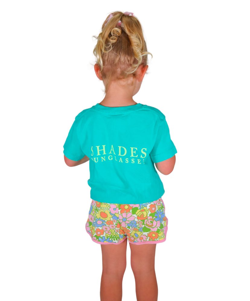 Child wearing a turquoise shirt with 'SHADES' branding and floral shorts on a white background
