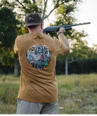 Man in a brown shirt with a duck and dog design holding a shotgun outdoors.