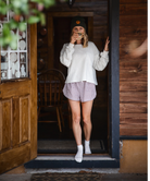 Person standing in a doorway wearing a white sweatshirt, checkered shorts, and a black cap.