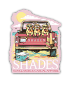 A die-cut sticker featuring a retro-style illustration of three golden retriever dogs wearing sunglasses in the bed of a pink pickup truck on a beach. The sticker includes the text 'SHADES' and 'SUNGLASSES & CASUAL APPAREL' on a white background.
