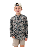A young boy smiling and wearing a long-sleeve camouflage hoodie with a beige cap and khaki shorts, photographed against a solid white background.