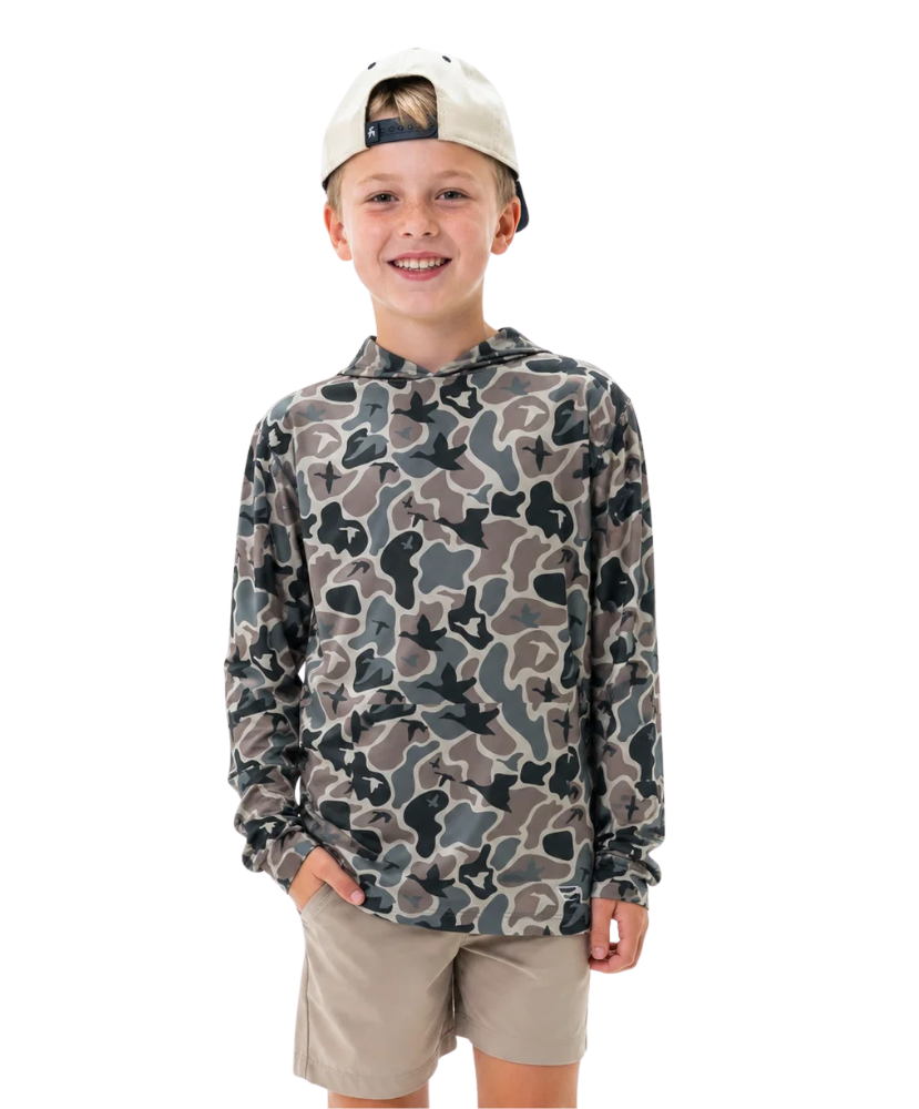 A young boy smiling and wearing a long-sleeve camouflage hoodie with a beige cap and khaki shorts, photographed against a solid white background.