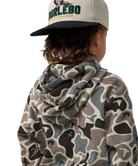 A child shown from the back wearing a beige and gray camouflage hoodie and a beige baseball cap with a black brim. The cap features the brand name 'BURLEBO OUTDOOR' in green and yellow.